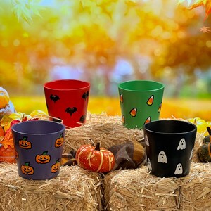 May include: Four painted terracotta flower pots with Halloween themes. The pots are decorated with bats, candy corn, ghosts, and pumpkins. The pots are sitting on a bed of straw.