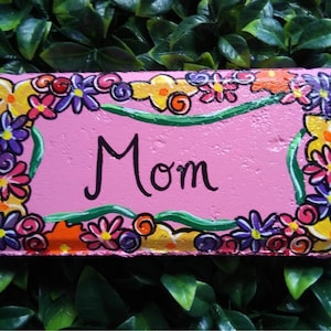 Könnte beinhalten: Ein rechteckiges, handbemaltes rosa Schild mit dem Wort "Mom" in schwarzer Schrift. Das Schild ist mit bunten Blumen in Gelb, Orange, Lila und Rosa sowie grünen Ranken verziert. Das Schild ist vor einem Hintergrund aus grünen Blättern platziert.
