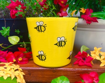 May include: Yellow ceramic planter with a hand-painted design of black bees. The planter is sitting on a wooden surface with green plants and red flowers.