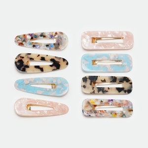 Puede incluir: Conjunto de ocho pinzas para el pelo de colores con un patrón de mármol. Las pinzas tienen forma ovalada y tienen una barra de metal dorado en el centro.