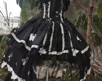 May include: Black Lolita dress with butterfly detailing. The dress has a fitted bodice with white lace trim and a layered skirt. White butterfly appliqués decorate the skirt. The dress is displayed on a mannequin.