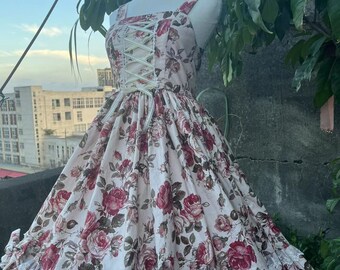 May include: A floral print dress with a square neckline and a corset-style bodice. The dress features a full skirt with a ruffled hem and a matching headband. The fabric is a light pink with red and pink roses.