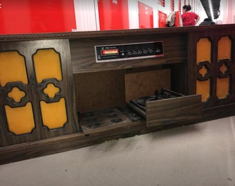 May include: A vintage wooden stereo console with a record player, a cassette player, and a radio. The console has a brown wood finish with yellow accents and a sliding drawer. The record player is visible with a turntable and a tone arm.