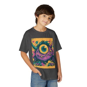 May include: A charcoal gray t-shirt featuring a colorful cartoon monster with one large eye, horns, and a purple body. The monster is set against a yellow background with colorful splatters. The shirt is worn by a person.