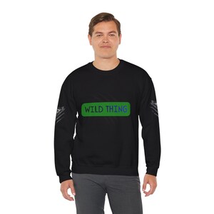 May include: A black long-sleeve sweatshirt featuring a green rectangular graphic on the chest with the text WILD THING printed in blue. The sleeves have a subtle grey patterned design on the outer sides, and the garment has a crew neckline.