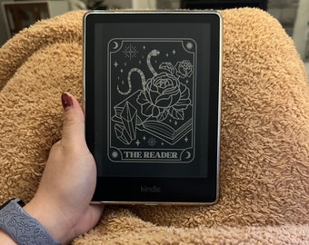 May include: A black and white illustration of a rose, snake, and crystal on a black Kindle e-reader. The text "THE READER" is displayed on the screen. The Kindle is on a brown fuzzy blanket.
