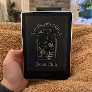 May include: A black and white Kindle e-reader displaying the text "The House of Wind Book Club" with a graphic of a moon, stars, and a staircase inside an arched window.