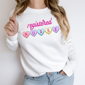 Könnte beinhalten: Weißes Sweatshirt mit dem Schriftzug "registered NURSE" in Pink und bunten herzförmigen Bonbon-Grafiken. Das Sweatshirt hat einen Rundhalsausschnitt und lange Ärmel.