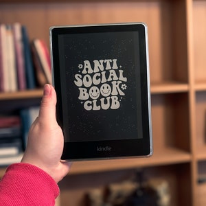 May include: A black Kindle e-reader displaying the text "ANTI SOCIAL BOOK CLUB" in a retro font with a smiley face. The e-reader is held in front of a bookshelf filled with books.
