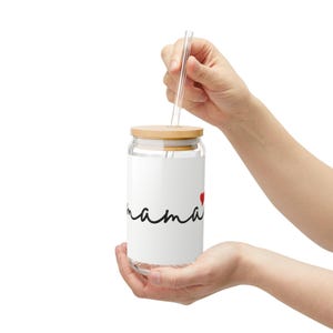 Puede incluir: Un vaso de vidrio transparente con tapa de madera y pajita transparente. El vaso tiene la palabra "mama" en escritura negra con un corazón rojo. El vaso está sostenido en las manos de una persona.