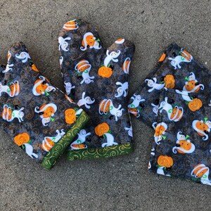 May include: Set of three Halloween-themed oven mitts and a pot holder. The fabric is black with a white ghost and orange pumpkin pattern. The mitts have green trim.