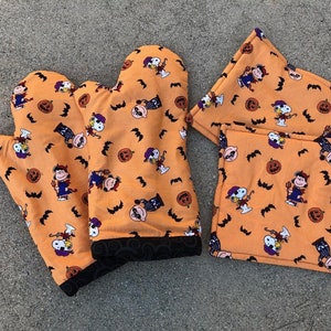 Może przedstawiać: Zestaw trzech pomarańczowych rękawic do piekarnika i podkładek na Halloween z powtarzającym się wzorem postaci z Peanuts przebranych w kostiumy na Halloween. Postacie to Snoopy, Woodstock i Linus. Rękawice i podkładki mają czarną lamówkę.