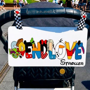 May include: A white rectangular stroller sign attached to a black stroller handle with checkered straps. The sign features the colorful text SPEND LOVE, with each letter styled after various cartoon characters, and the word STROLLER printed below.