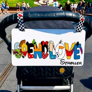 May include: A white stroller sign with the word "SEND LOVE" in colorful, cartoon-style letters. The sign is attached to a black stroller with a black handle. The background shows a sunny day at a theme park.