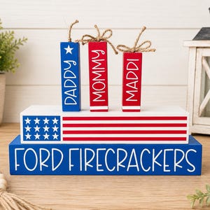 Puede incluir: Una exhibición patriótica de madera con una base azul con la inscripción "FORD FIRECRACKERS". Encima, un bloque blanco con un diseño de rayas rojas y blancas y un campo de estrellas azules. Tres adornos de petardos de madera están etiquetados como "DADDY", "MOMMY" y "MADDI".