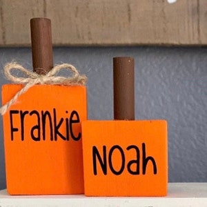 May include: Two orange wooden pumpkins with the names Frankie and Noah painted on them. The pumpkins are decorated with twine bows.