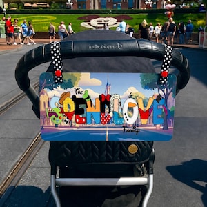 May include: A stroller with a custom sign that reads "THE END LOVE Family" in colorful, cartoon-style lettering. The sign features Disney characters and is attached to the stroller handle with polka dot straps.