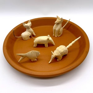 May include: Five wooden animal figurines on a terracotta plate. The figurines are a cat, a fish, an armadillo, a pig, and a fox. The figurines are all carved in a simple style and are painted in a natural wood color.