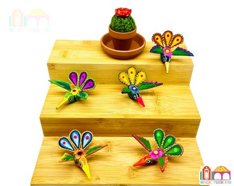 Pode incluir: Cinco beija-flores de madeira coloridos com detalhes pintados, dispostos em uma tela de madeira de vários níveis. Os beija-flores têm acentos verdes, amarelos, azuis e rosados. Um pequeno cacto em vaso com flores vermelhas está no nível superior.