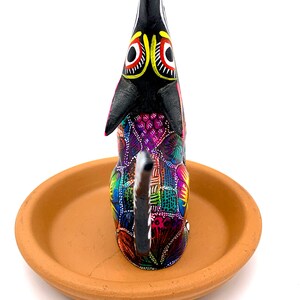 May include: A black and colorful wooden dog figurine with a clay base. The dog is painted with intricate patterns in shades of pink, purple, blue, green, yellow, and orange. The dog's eyes are black with yellow and red accents.