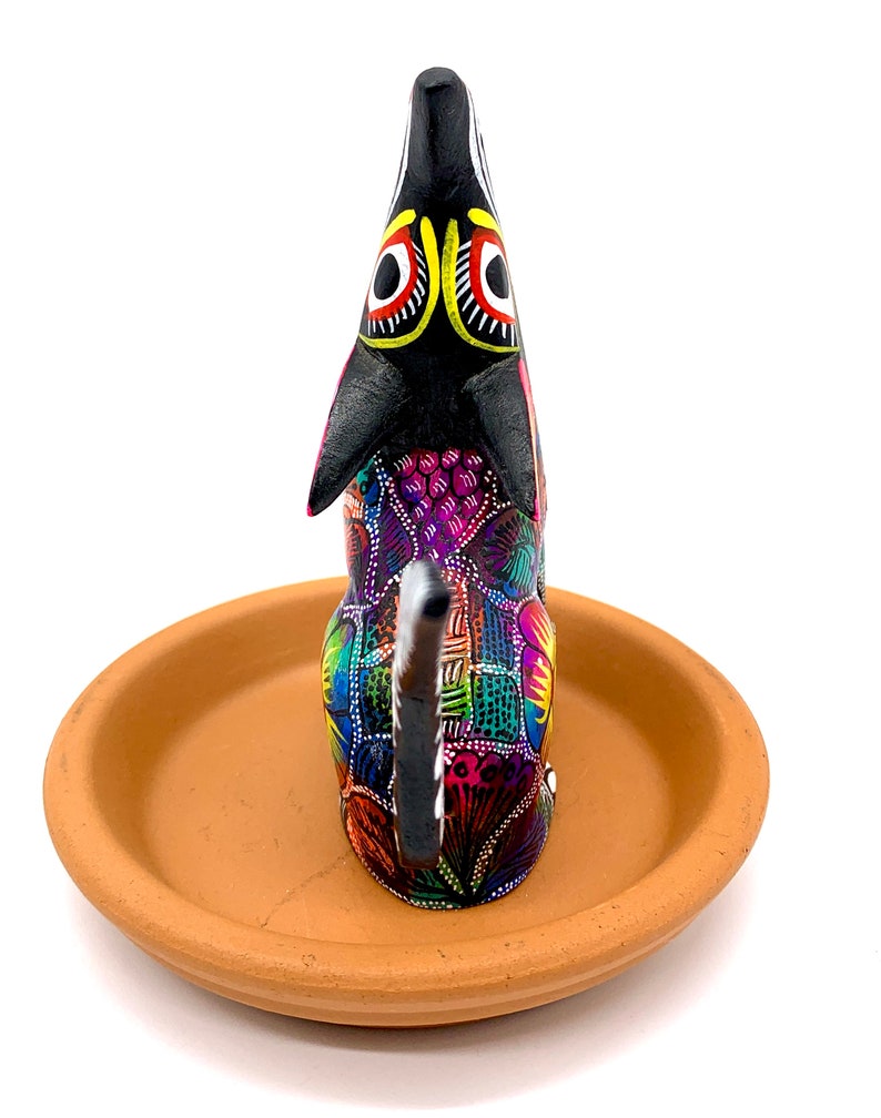 May include: A black and colorful wooden dog figurine with a clay base. The dog is painted with intricate patterns in shades of pink, purple, blue, green, yellow, and orange. The dog's eyes are black with yellow and red accents.