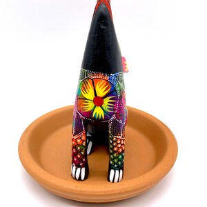 May include: A black wooden figurine of a dog with colorful floral patterns and white paws. The dog is standing on a terracotta saucer.