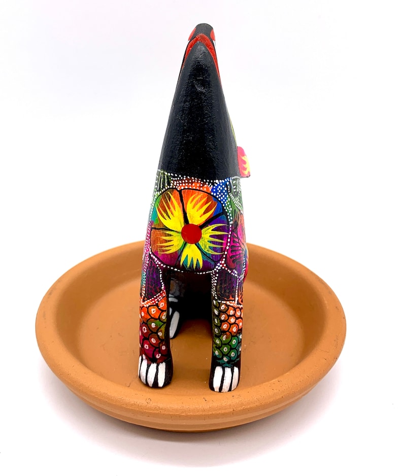 May include: A black wooden figurine of a dog with colorful floral patterns and white paws. The dog is standing on a terracotta saucer.
