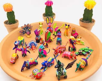 May include: A collection of colorful, hand-painted wooden animal figurines, including a peacock, a unicorn, a spider, and various other creatures. The figurines are arranged on a terracotta plate, surrounded by two potted cacti.