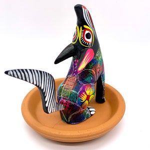 May include: A black wooden coyote figurine with colorful painted details, including flowers and geometric patterns. The coyote is standing on a terracotta dish.