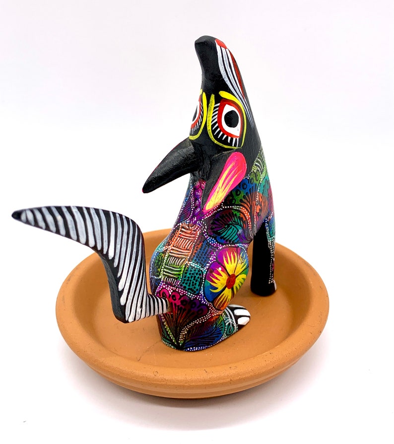 May include: A black wooden coyote figurine with colorful painted details, including flowers and geometric patterns. The coyote is standing on a terracotta dish.