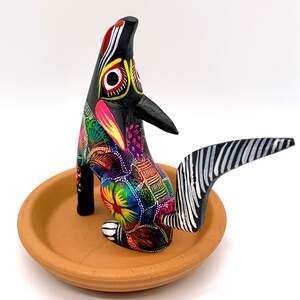 May include: A black wooden animal figurine with colorful painted details, standing on a terracotta dish. The animal has a long, striped tail and is painted with a variety of colors, including red, yellow, green, blue, and purple.