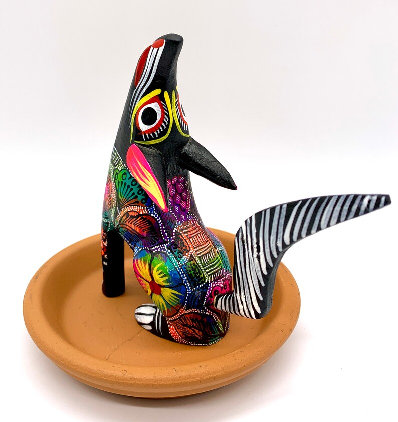May include: A black wooden animal figurine with colorful painted details, standing on a terracotta dish. The animal has a long, striped tail and is painted with a variety of colors, including red, yellow, green, blue, and purple.