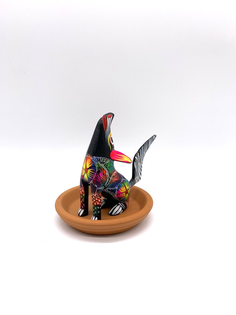 May include: A black wooden figurine of a dog with colorful floral patterns painted on its body. The dog is sitting on a small brown clay dish.