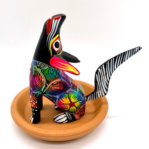 May include: A hand-painted wooden figurine of a dog with a black and white striped tail. The dog is painted in bright colors, including pink, yellow, green, blue, and orange. The dog is sitting on a terracotta-colored base.