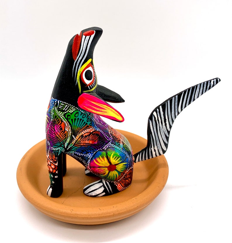 May include: A hand-painted wooden figurine of a dog with a black and white striped tail. The dog is painted in bright colors, including pink, yellow, green, blue, and orange. The dog is sitting on a terracotta-colored base.