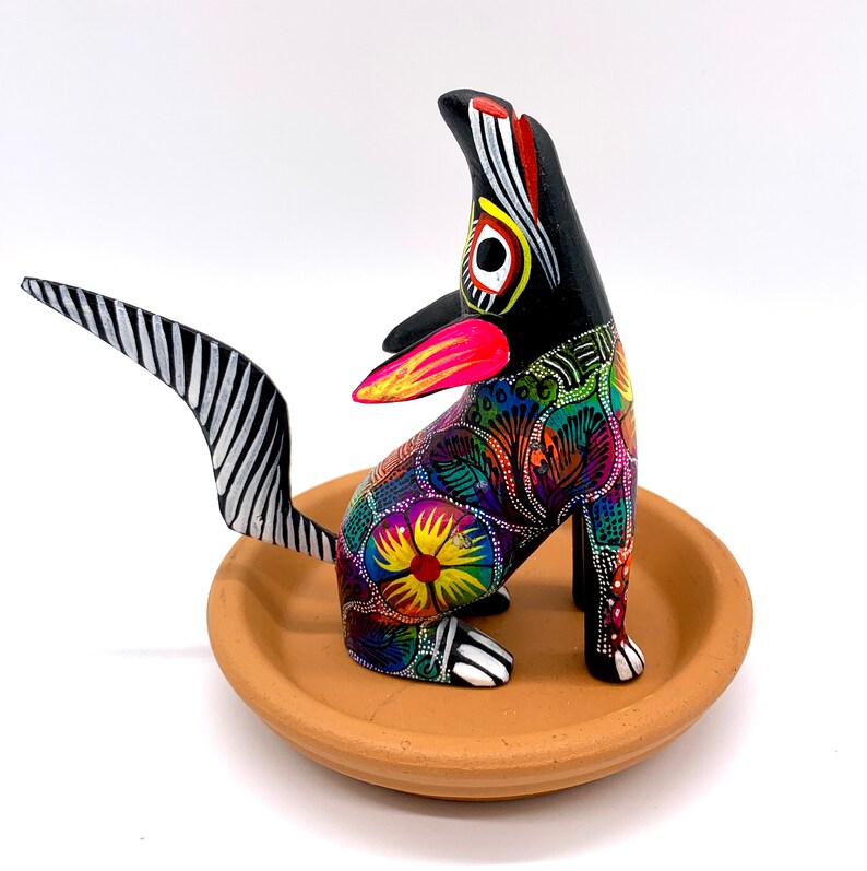May include: A colorful, hand-painted wooden dog figurine with black and white stripes on its tail. The dog is sitting on a terracotta-colored base and has a bright pink tongue sticking out.
