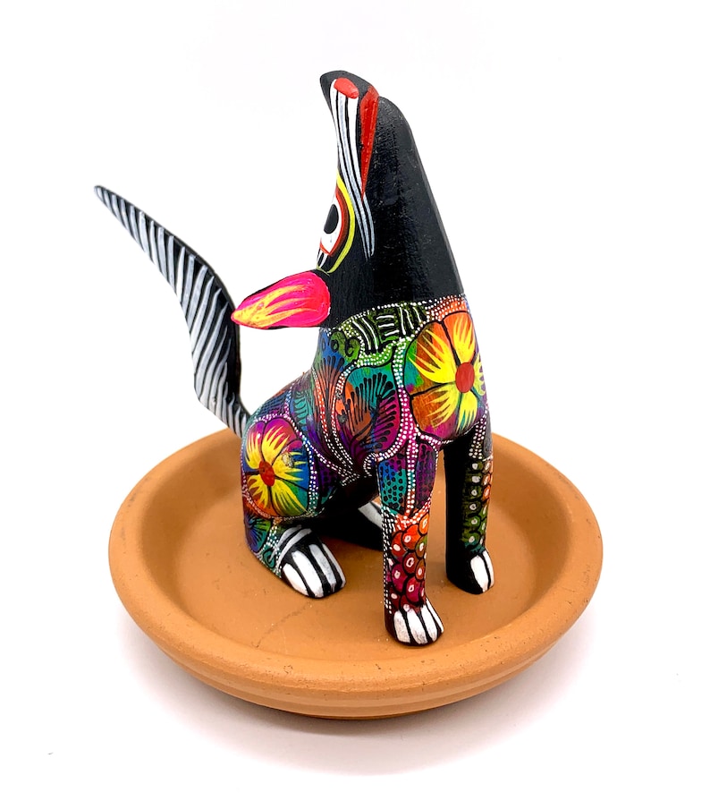 May include: A black wooden dog figurine with colorful floral patterns and a white and black striped tail. The dog is sitting on a terracotta clay dish.