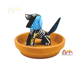 May include: A black and white dog figurine with blue and yellow details, sitting in a terracotta dish. The dog is a traditional Mexican folk art piece.