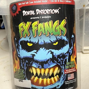 Może przedstawiać: Akcesorium kostiumu na Halloween, FX Fangs, w opakowaniu. Opakowanie przedstawia niebieską twarz potwora z żółtymi oczami i zestawem żółtych kłów. Tekst na opakowaniu brzmi "Dental Distortions" i "Take Your Halloween To The Extreme!"