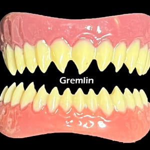 May include: A set of novelty teeth with yellow fangs and pink gums. The word "Gremlin" is printed in white text below the top set of teeth. The teeth are set against a black background.