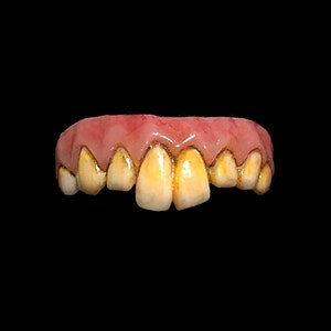 May include: A set of yellowed, crooked false teeth with a pink gum line. The teeth are discolored and appear to be aged, with a slightly uneven arrangement. The background is solid black.