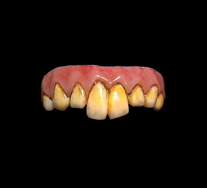 May include: A set of yellowed, crooked false teeth with a pink gum line. The teeth are discolored and appear to be aged, with a slightly uneven arrangement. The background is solid black.