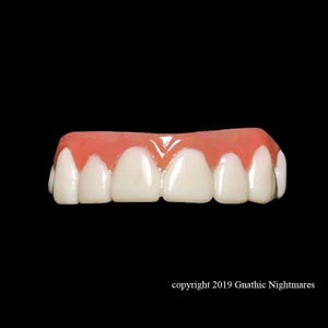 May include: A set of upper teeth with white enamel and pink gums. The teeth are arranged in a row, with a slight curve. The background is solid black. The text "copyright 2019 Gnathic Nightmares" is at the bottom.