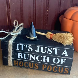 Peut inclure: Blocs de bois noirs empilés avec le texte "IT'S JUST A BUNCH OF HOCUS POCUS". Un chapeau de sorcière noir miniature et un balai sont posés dessus, attachés avec de la ficelle. Une citrouille orange est en arrière-plan.