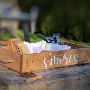 May include: A wooden box with the word "s'mores" written on the front. The box is filled with graham crackers, chocolate bars, and marshmallows.