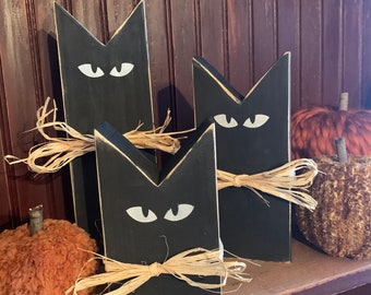 May include: Three black wooden cat figurines with white painted eyes and straw accents. The cats are arranged in a row on a wooden shelf with a brown and orange pumpkin beside them.