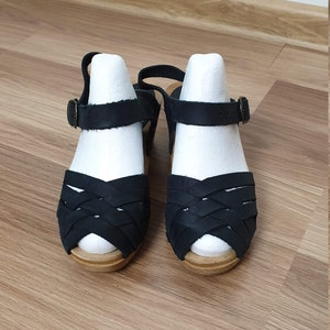 Op de afbeelding: Een paar zwarte leren sandalen met een geweven ontwerp en houten zolen.