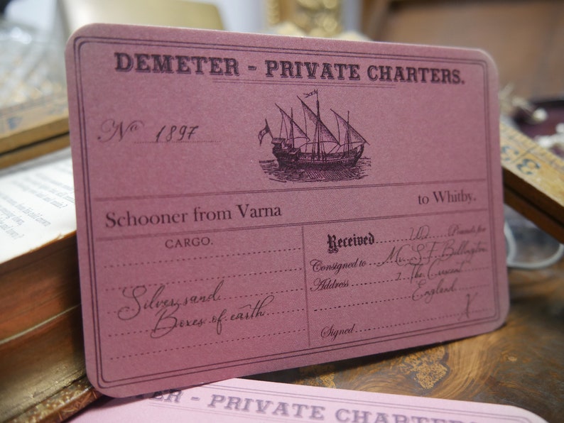 May include: A vintage pink card with the text "DEMETER - PRIVATE CHARTERS."  The card has a handwritten number "N 1897" and a black and white illustration of a sailing ship. The text "Schooner from Varna to Whitby" is printed on the card. The card has a section for cargo and a section for receiving the cargo. The cargo section lists "Silvery sand Boxes of earth."