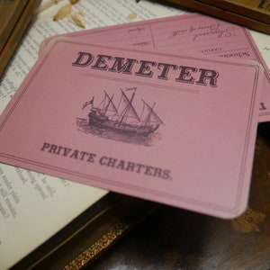 May include: A pink card with the text "DEMETER PRIVATE CHARTERS."  A black and white illustration of a sailing ship is in the center of the card.