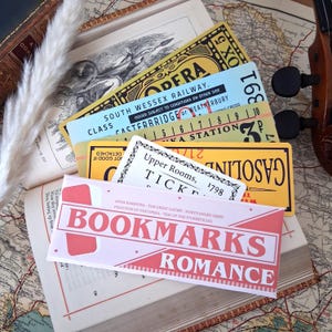 May include: A collection of rectangular paper bookmarks featuring vintage-style ticket designs in yellow, light blue, and white. The front bookmark is pink and white with the text BOOKMARKS ROMANCE and a list of classic novel titles printed on it.